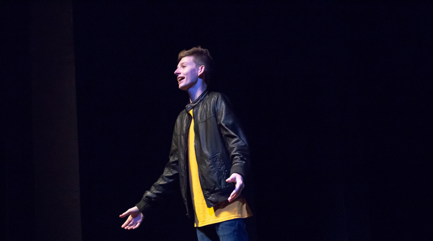 student wearing black leather jacked and yellow shirt singing on stage