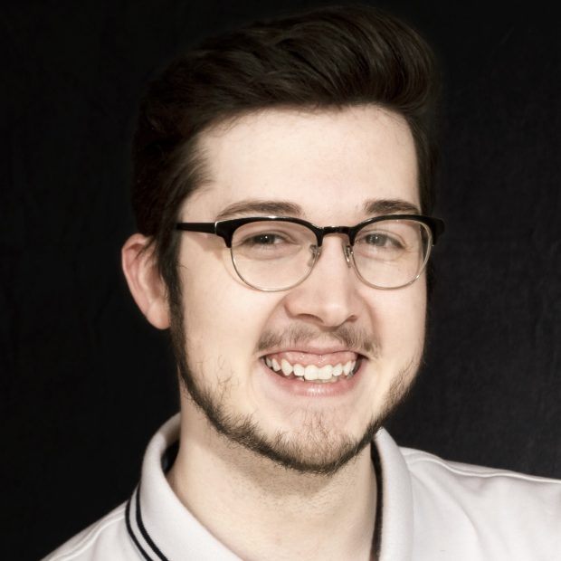 Man with glasses with brown hair and a beard with a white collared shirt and a dark background