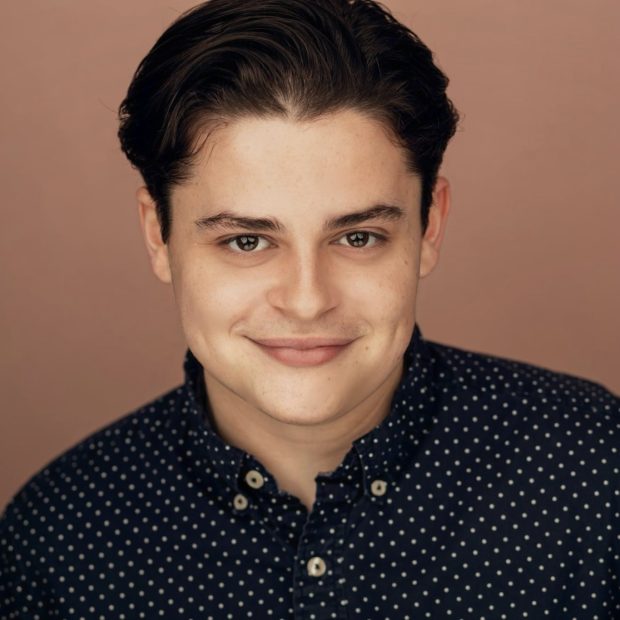 Young man with brown hair and eyes in a navy and white polka dot patterned shirt with a brown background